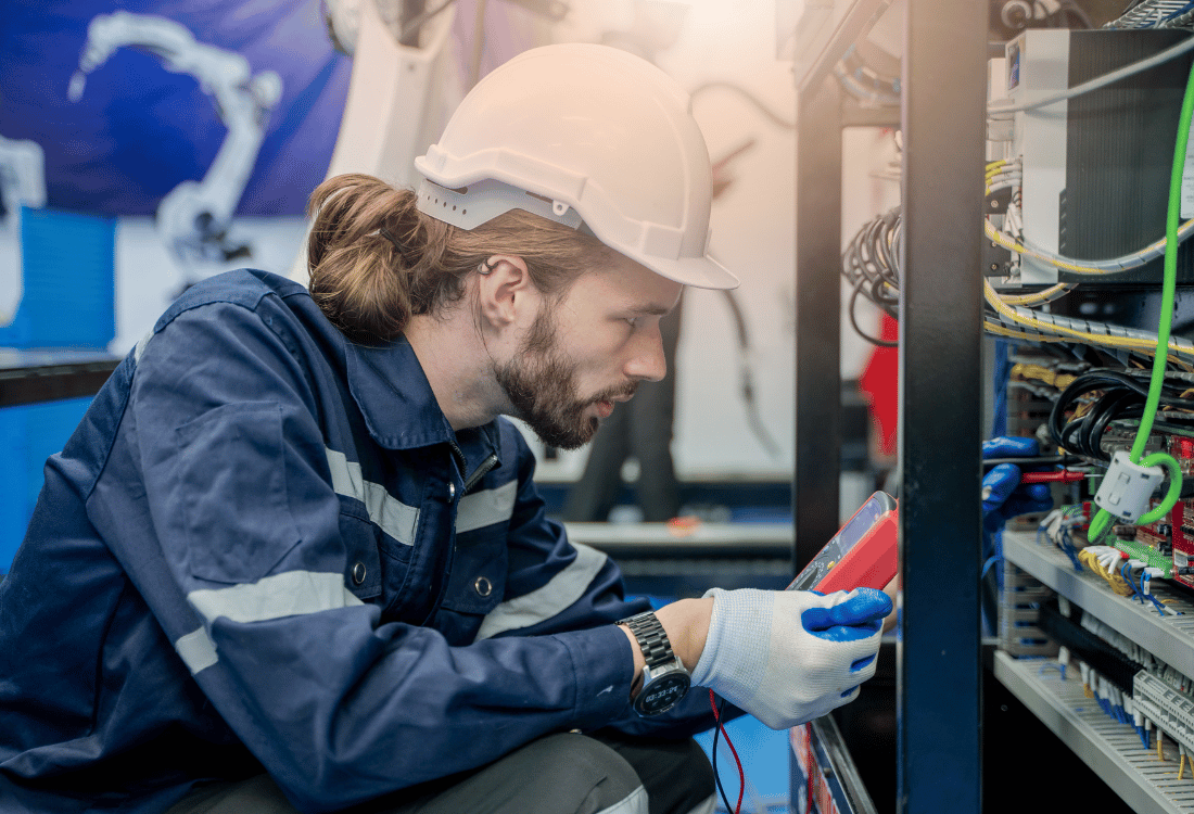 A maintenance engineer troubleshooting with a handheld device connected to a network panel, ensuring operational efficiency and continuity of their organisation's production output as part of the wider supply chain.